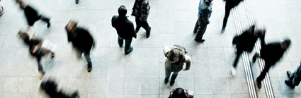 Several individuals walking swiftly on a grey concrete ground whist four individuals stand stationary, scattered amongst the rest.