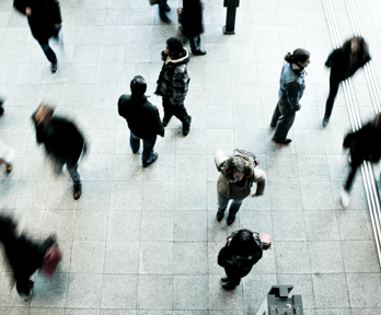 Several individuals walking swiftly on a grey concrete ground whist four individuals stand stationary, scattered amongst the rest.