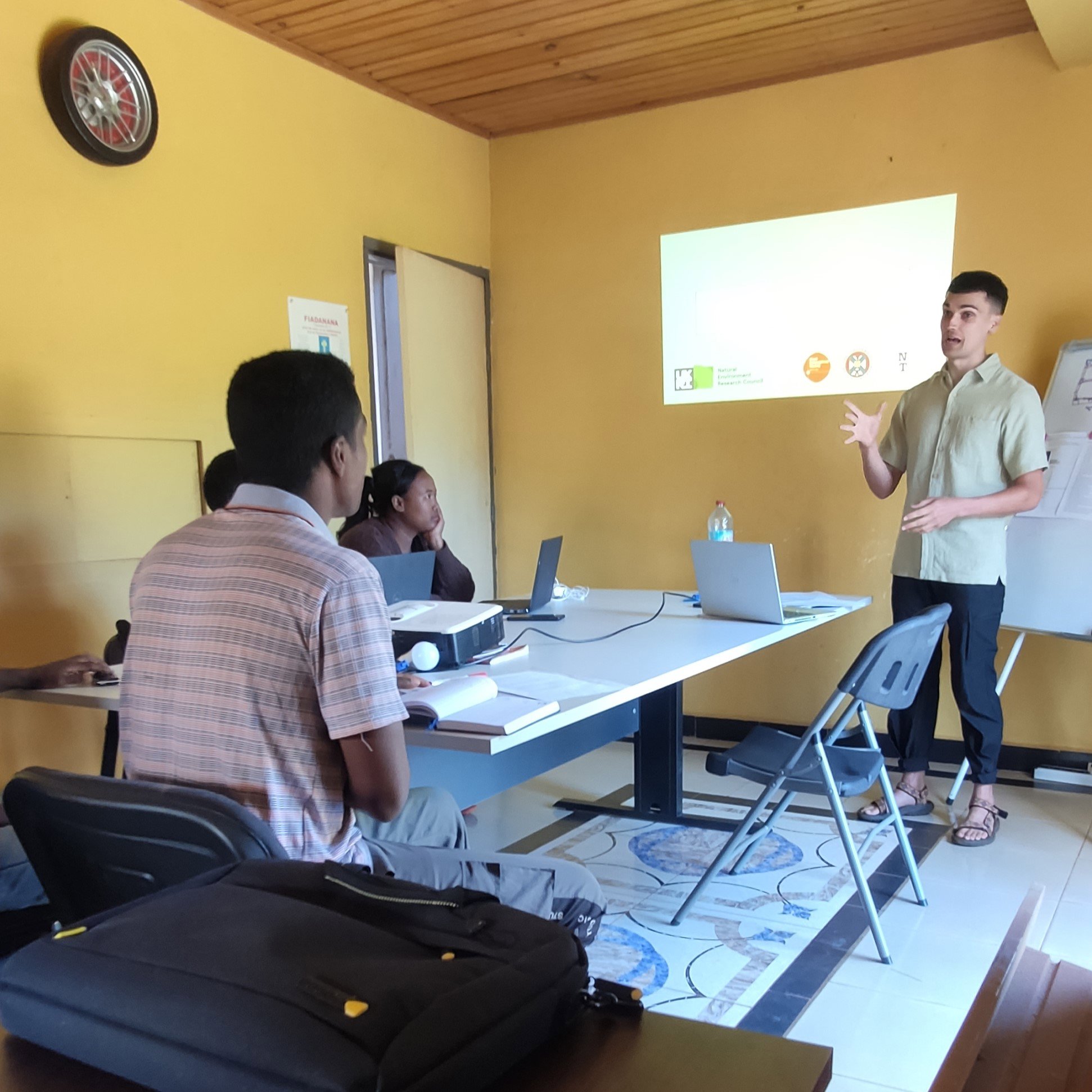 Person stood up giving a presentation to three people sat around a table. A digital projector is projecting a presentation on the wall. 