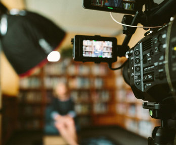 A person sitting in front of a bookcase and a camera set up for an interview.