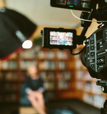 A person sitting in front of a bookcase and a camera set up for an interview.