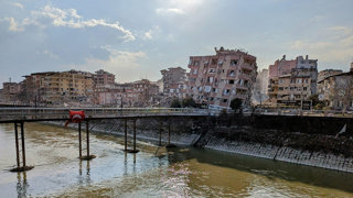 Damaged appartment buildings at the bring of toppling over. A river and bridge are in the foreground, with over the bridge a Turkish flag.