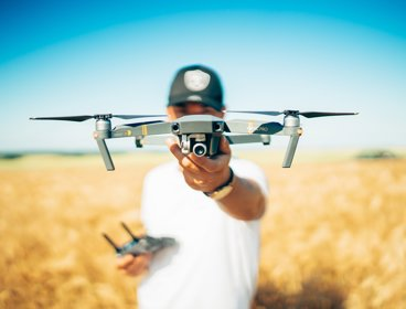 A man standing in yellow field of wheat, holding a silver drone out towards the camera. The drone obscures his face.