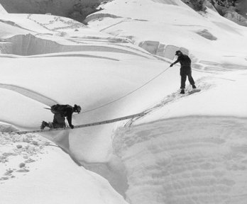 Black and white photo of two explorer in a desolate, snowy area crossing a gap using a ladder horizontally.