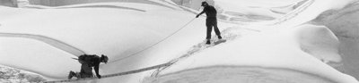 Black and white photo of two explorer in a desolate, snowy area crossing a gap using a ladder horizontally.