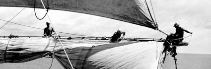 Old, monochrome photograph of people hoisting the sails of a sailing ship.