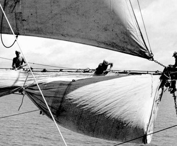 Old, monochrome photograph of people hoisting the sails of a sailing ship.