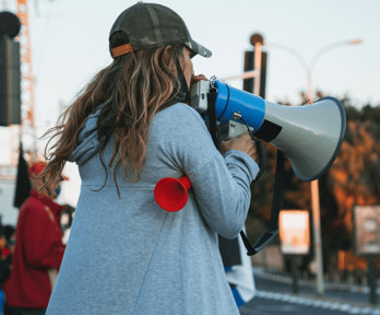 Person speaking into a microphone with a blurred background showing other protesters