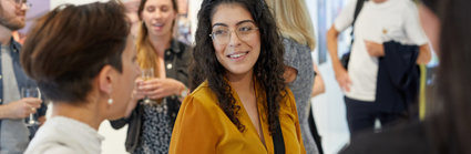 A woman with long dark hair in a mustard coloured blouse stand amongst a group of people chatting and drinking in an exhibition space.