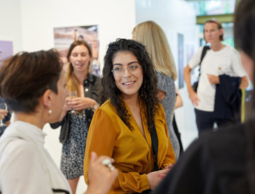 A woman with long dark hair in a mustard coloured blouse stand amongst a group of people chatting and drinking in an exhibition space.