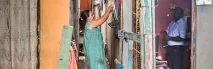A person hangs up wet clothes on a washing line outside of the house whilst a person on the other side of the wall, inside the house stands and stares at the camera.