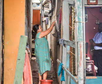 A person hangs up wet clothes on a washing line outside of the house whilst a person on the other side of the wall, inside the house stands and stares at the camera.