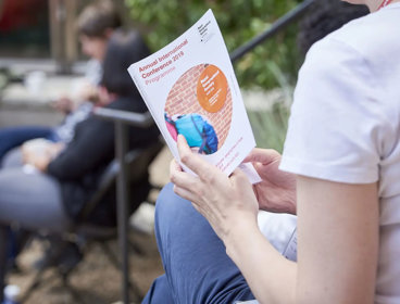 A person sat down outside holding a borchure. The front page reads 'Annual International Conference 2019 Programme'.