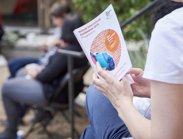 A person sat down outside holding a book. The front page reads 'Annual International Conference 2019 Programme'.