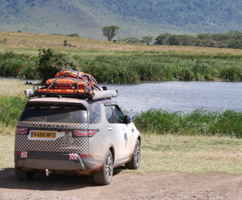 A car with equipment attached to the roof parked in front of lake. There is vegetation surrounding the bank of the lake with a row of trees behind.