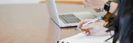 Two people sit next to each other at a wooden table. one sits before a notebook and holds a pen whilst the other sits in front of a laptop