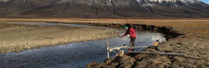 A researcher collecting water temperature measurements at the edge of a river in a baron landscape, with mountains in the background. 