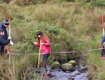 Three teachers in a grassy field assist a student, who is standing in a small stream, carrying out fieldwork research.