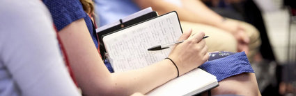 Audience member with a notebook and pen on their lap, seated next to other people.