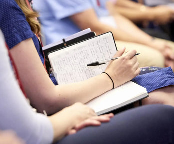 Audience member with a notebook and pen on their lap, seated next to other people.