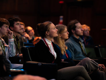 A white blond woman looks at the stage while surrounded by a small group of audience members of mixed genders sitting in the Society's lecture theatre.
