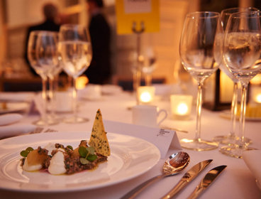 A formal dinner table with food, cutlery, wine glasses and glass candleholders with lit tealights.