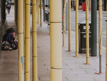 A person sitting on pavement which features scaffolding, a black bin and a bus stop.  The image suggests this person is homeless.