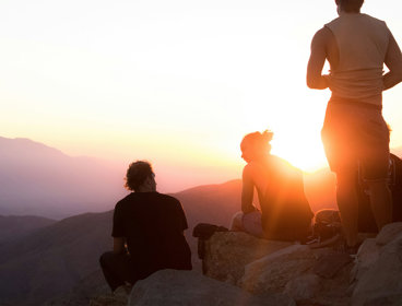 Two people sitting and one person standing near cliff taken during golden hour.