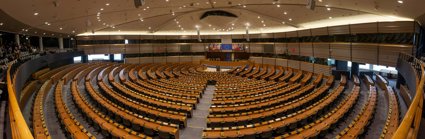 Circular rows of seats in a conference room with a stage at the front. There are European flags behind the stage. 