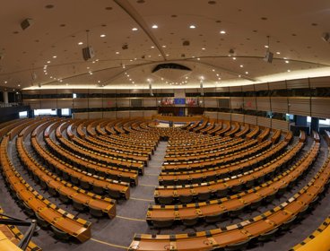 Circular rows of seats in a conference room with a stage at the front. There are European flags behind the stage. 