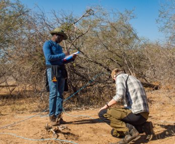 Two researchers carrying out an investigation in an arid landscape. One researcher is standing holding a notebook and writing notes, and the other is crouched on the ground. 
