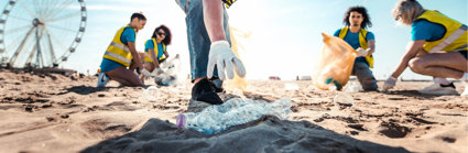 People picking up litter on a beach on a sunny day. In focus is a person reaching for a plastic bottle.