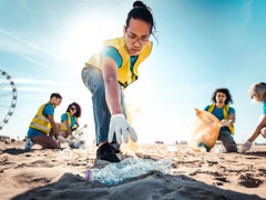 People picking up litter on a beach on a sunny day. In focus is a person reaching for a plastic bottle.