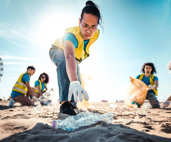 People picking up litter on a beach on a sunny day. In focus is a person reaching for a plastic bottle.