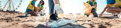 People picking up litter on a beach on a sunny day. In focus is a person reaching for a plastic bottle.