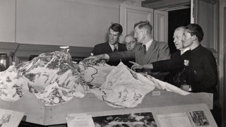 Black and white photo of the Everest plaster model with Edmund Hillary, Wordie, John Hunt, Herbert and Tenzing Norgay viewing and pointing at the model.