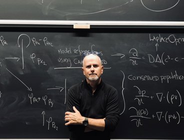 Photograph of John Hessler standing in front of a blackboard with equations written in chalk