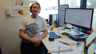 A researcher sitting at a desk in his office with his research showing on the monitor next to him.