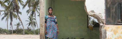 A local resident of Fuvemeh, Ghana, standing on a beach while leaning against the wall of a building damaged by coastal erosion.