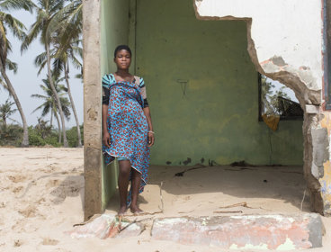 A local resident of Fuvemeh, Ghana, standing on a beach while leaning against the wall of a building damaged by coastal erosion.