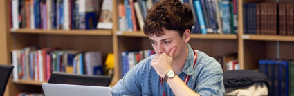 Person sat at a table looking at a laptop screen with notepads and books surrounding the laptop. Behind the table there are bookshelves with stacks of books. 