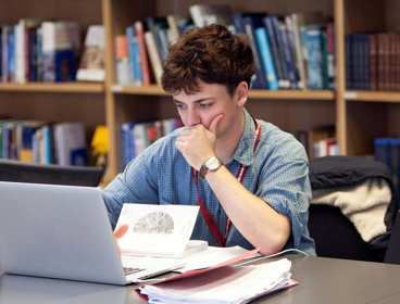 Person sat at a table looking at a laptop screen with notepads and books surrounding the laptop. Behind the table there are bookshelves with stacks of books. 