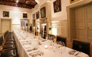 A long dinner table with wooden chairs and white linen setup down the centre of a grand hall.