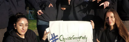 A group of students are standing holding a sign that reads 'Choose Geography'. They are pointing at it and smiling