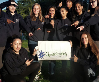 A group of students are standing holding a sign that reads 'Choose Geography'. They are pointing at it and smiling