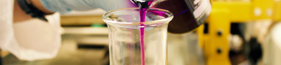 A person wearing a lab coat and blue gloves pours a purple liquid form one beaker into another
