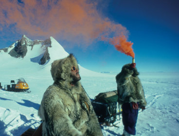 Two people standing in the snow, one holding a flare above their head. 