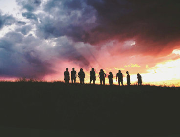 Silhouette of nine people standing on a hill against a sunrise.