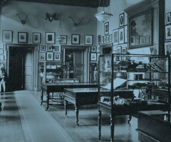 Old black and white photograph of the Geographical Museum showing multiple display cases, paintings on the walls, and a large globe.