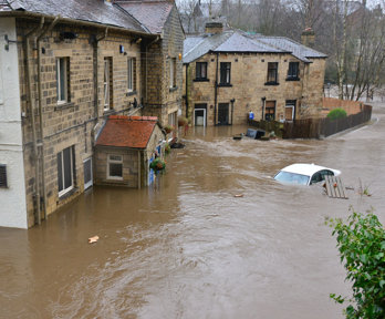 a residential area where flood water has risen; the flood water is almost reaching the downstairs floor windows and a white car is almost fully submerged.